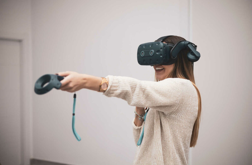 A young woman is smiling while wearing a virtual reality headset, and a holding out her left hand using a virtual reality motion controller.