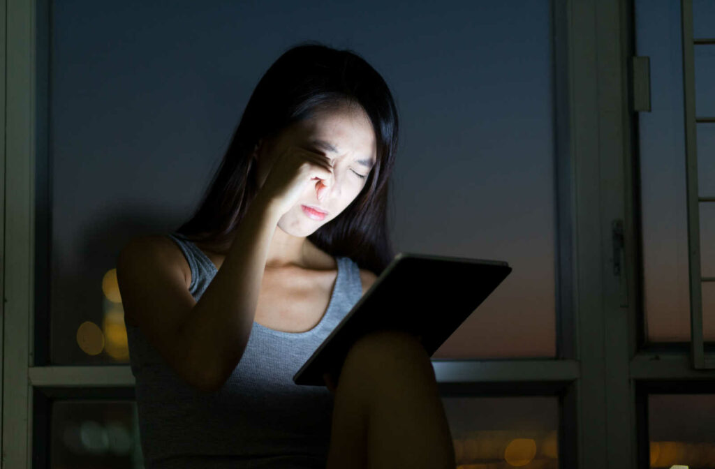 A young Asian woman sitting at a window sill in the dark. She is holding up a glowing iPad to her face with her right hand, and rubbing her eyes with her left hand while both eyes are closed.