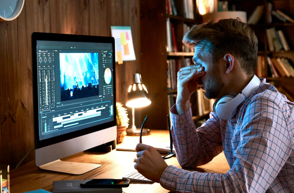 A young man is massaging his eyes while working on his computer. His eyes are sensitive to light due to a lack of sleep.